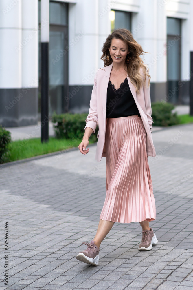 a young girl walks along the sidewalk in wide strides. Skirt plise, long jacket, sneakers. Long wavy hair. Very beautiful European in the summer walks down the street in a great mood.