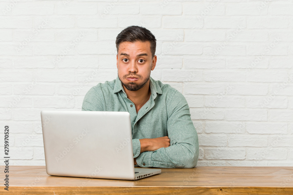 Young filipino man sitting working with his laptop blows cheeks, has tired expression. Facial expression concept. Young filipino man sitting working with his laptop blows cheeks, has tired expression. Facial expression concept.