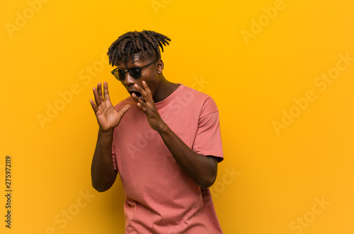 Young casual black man wearing sunglasses shouts loud, keeps eyes opened and hands tense.
