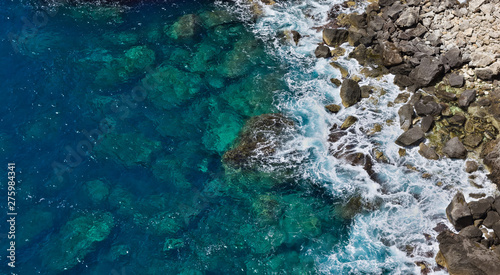 Aerial top view of crystal clear blue waves hitting rocky beach creating white foam.