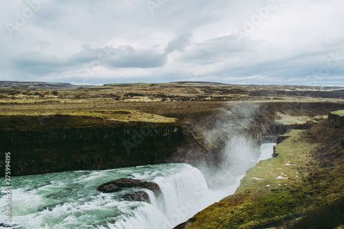 Green fields, black rocks, and turquoise water of Gullfoss waterfall in Iceland. Clouds of splashes flying in the air