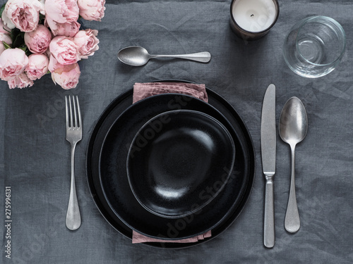 Beautiful table setting on gray linen tablecloth. Festive table setting for wedding dinner with pink spray roses and scented cabdle. Holiday dinner with black plates. Dinner, utensil. Copy space
