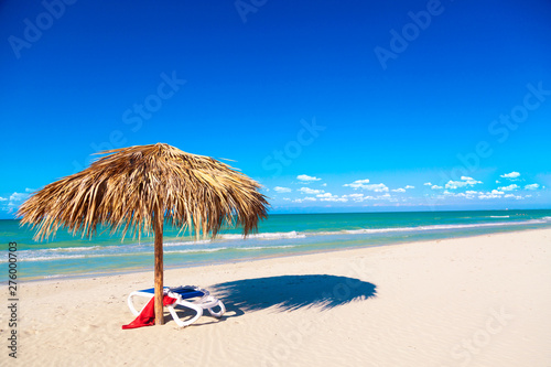 Fototapeta Naklejka Na Ścianę i Meble -  A sun lounger with red towel under an umbrella on the sandy beach by the sea and cloudy sky. Vacation background. Idyllic beach landscape. Free space for your text