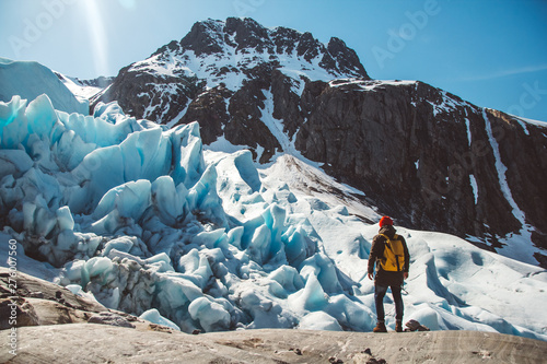 Traveler man with a yellow backpack wearing a red hat standing on a rock on the background of a glacier, mountains and snow. Travel lifestyle concept. Shoot from the back