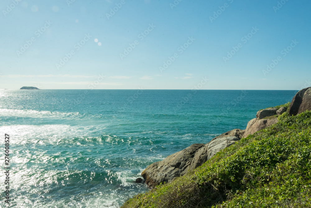Praia com rochas e vegetação banhada por um mar azul em um dia de Sol