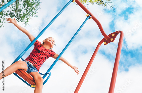 Funny smiling little girl swinging on the swing wide opened an arms with blue sky background