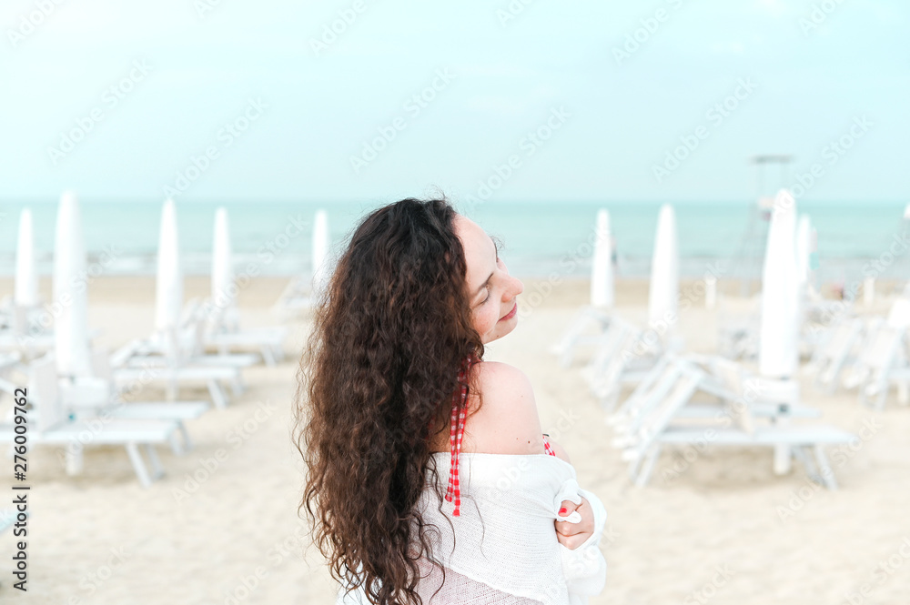 A woman in a red dress and curly hair is standing on the beach. A man with happy emotions. Walk along the sea promenade. Travel and tourism in the summer. Copy space