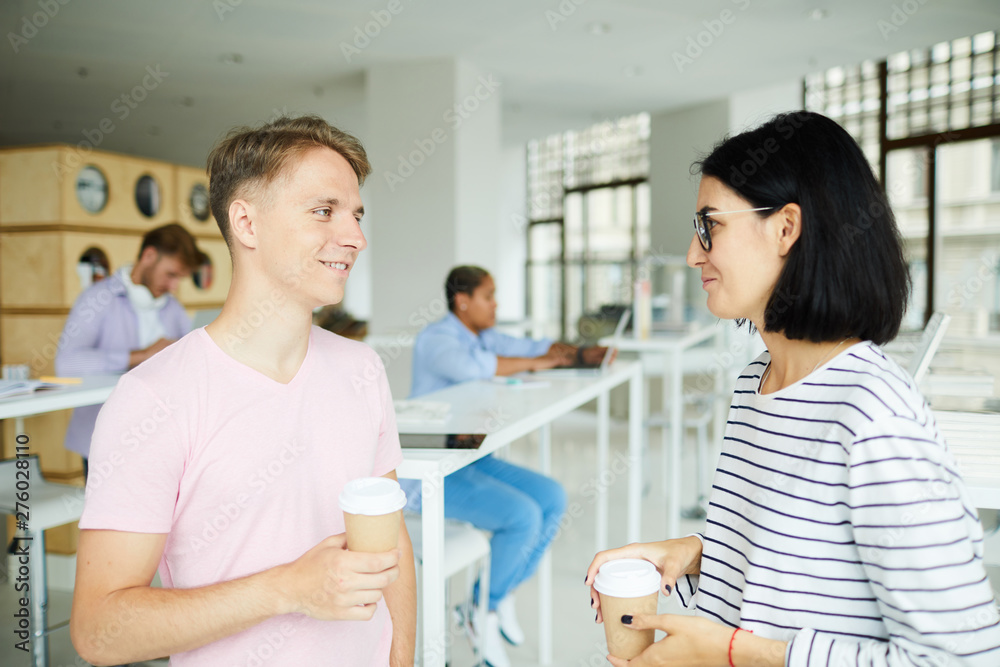 Positive confident young students in casual wear standing in campus hall and drinking coffee while discussing joint project Positive confident young students in casual wear standing in campus hall and drinking coffee while discussing joint project