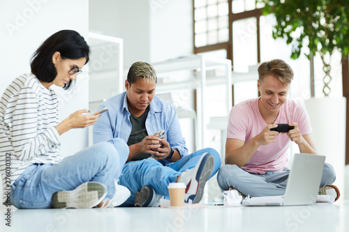 Group of technology-addicted students in casual clothing sitting in relaxed poses on floor and using modern devices while searching for information in internet