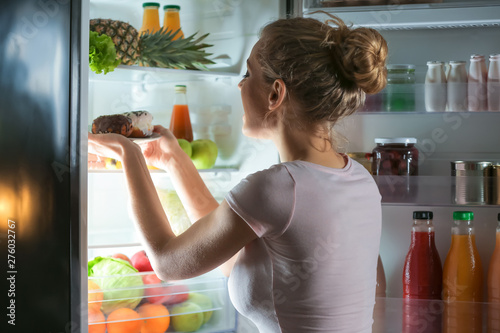 Fotografia Beautiful young woman choosing food in refrigerator at night