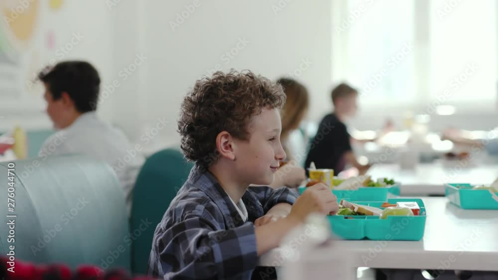 Happy little pupils having a lovely break during classes at school. View of primary students eating lunch from containers talking and sitting in the school canteen.