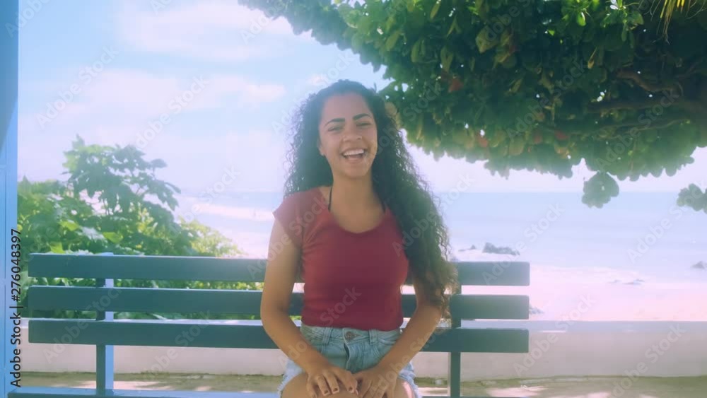 Woman siting on the bench. Beautiful young woman with curly hair smiling with the sea in the background.