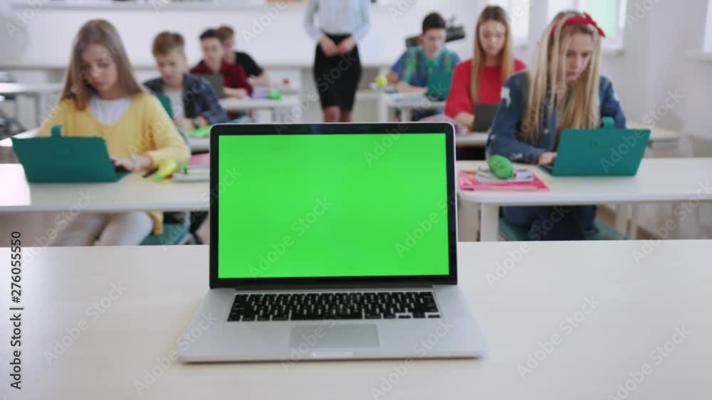 Computer science lesson in a modern school. New modern opened computer laptop with green screen on a teacher board. Female educator walking between students watching them studying online.