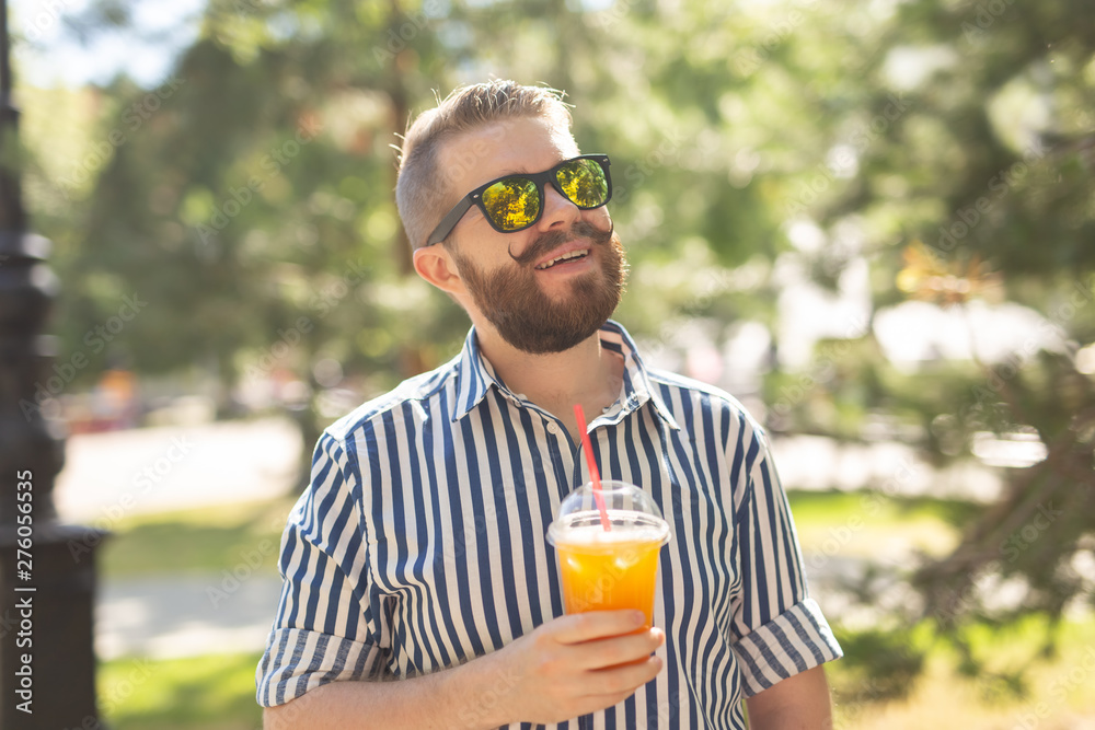 Portrait of a positive cheerful young man with a glass of juice with a straw while walking in the park on a warm sunny summer day. The concept of rest after study and work on weekends. Portrait of a positive cheerful young man with a glass of juice with a straw while walking in the park on a warm sunny summer day. The concept of rest after study and work on weekends.