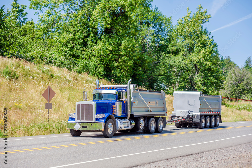 Blue big rig semi truck tipper with two tip trailers going on the road with hill