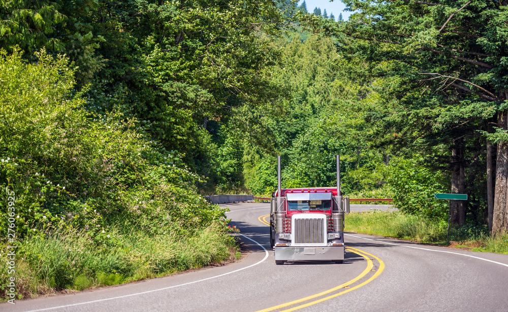 Red big rig classic American semi truck with flat bed semi trailer running on the winding road in green forest