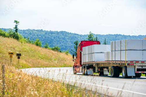 Red big rig semi truck transporting packed and tightened with slings lumber on flat bed semi trailer running downhill on the winding road photo