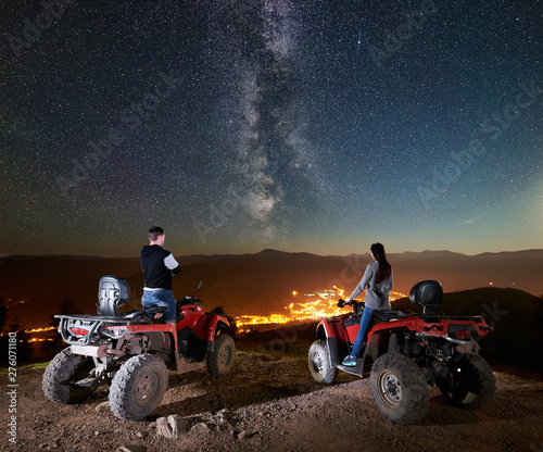 Rear view of happy couple man and woman tourists standing on atv quad motorbikes on the top of mountain, enjoying beautiful view of night sky full of stars, Milky way, luminous village on background