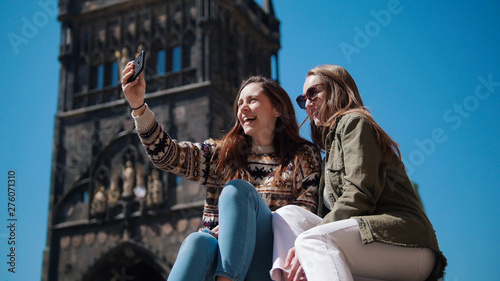 Two young smiling women sit under the old tower and take a selfie photo