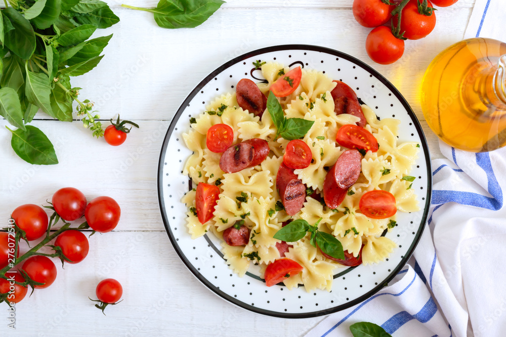 Tasty pasta farfalle with grilled sausages, fresh cherry tomatoes and basil on a plate on a white wooden background. Top view, flat lay. Tasty pasta farfalle with grilled sausages, fresh cherry tomatoes and basil on a plate on a white wooden background. Top view, flat lay.