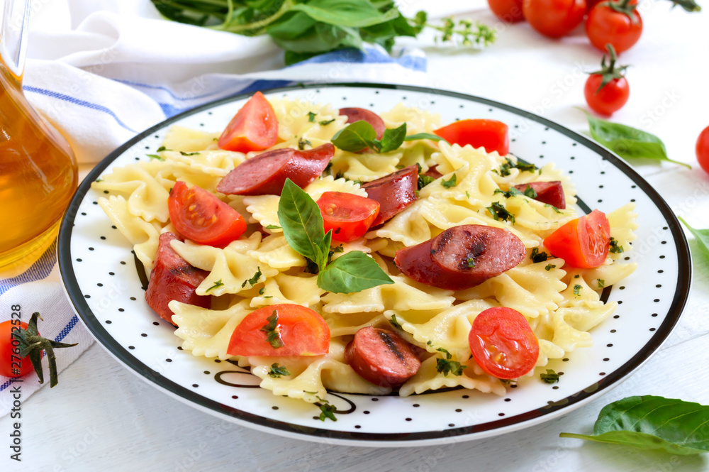Tasty pasta farfalle with grilled sausages, fresh cherry tomatoes and basil on a plate on a white wooden background.