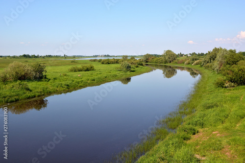 Summer rural landscape with a calm river with a reflection of trees and blue sky in the water with green fields with trees and a village on the banks.River Veriaja Novgorod region