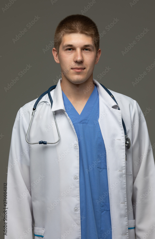 Closeup shot of young caucasian man doctor standing with crossed arms in white uniform against grey background looking perfectly confident.