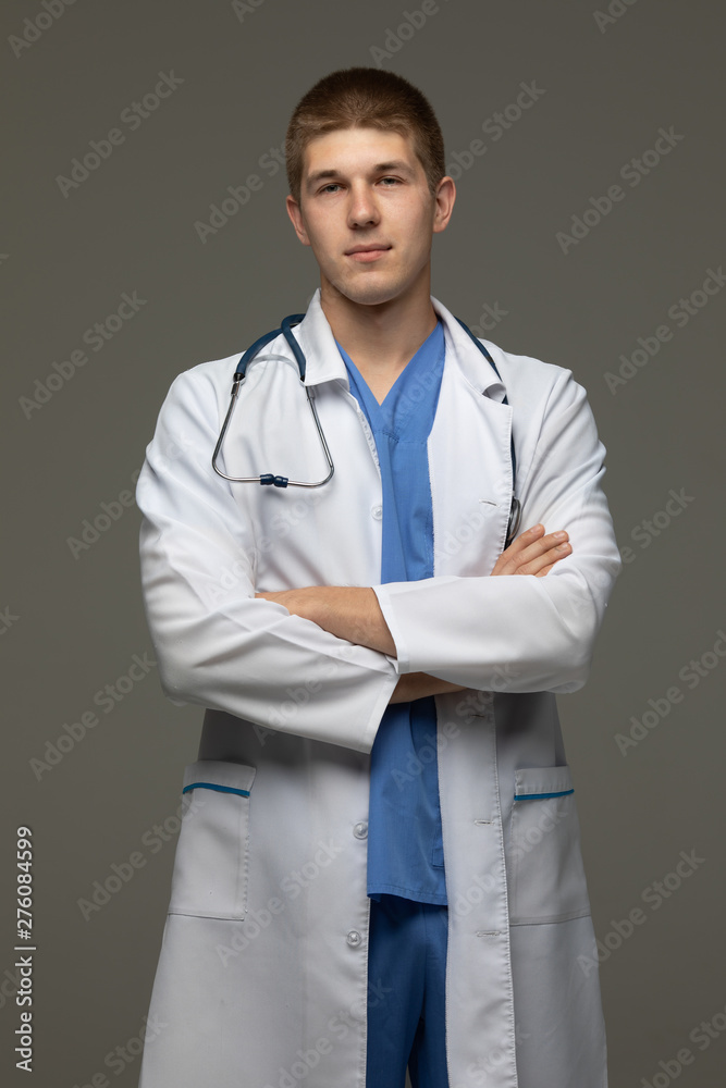 Closeup shot of young caucasian man doctor standing with crossed arms in white uniform against grey background looking perfectly confident. Closeup shot of young caucasian man doctor standing with crossed arms in white uniform against grey background looking perfectly confident.