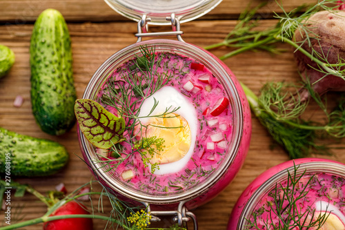 Cold beetroot soup, cold served with the addition of cucumbers, radishes, boiled eggs and fresh herbs, dill, green onions in glass jars on a wooden table, top view. Chilled Lithuanian cold soup  