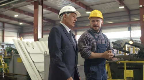 Tracking medium shot of mature factory executive walking along sunlit metalworking facility being briefed by smiling engineer, with racks of metal components and equipment in background photo