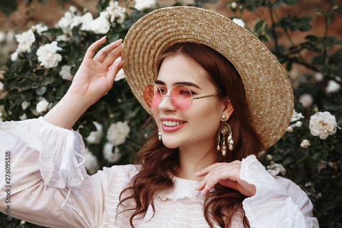 Outdoor close up portrait of young beautiful happy smiling lady wearing straw hat, pink round sunglasses, vintage pearl earrings, model looking aside