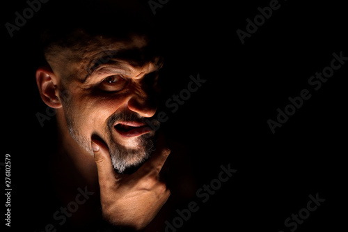  Face with a bearded man grimace against a dark background with sharp shadows. Comedic, fabulous villain or negative character conception with copy space.
