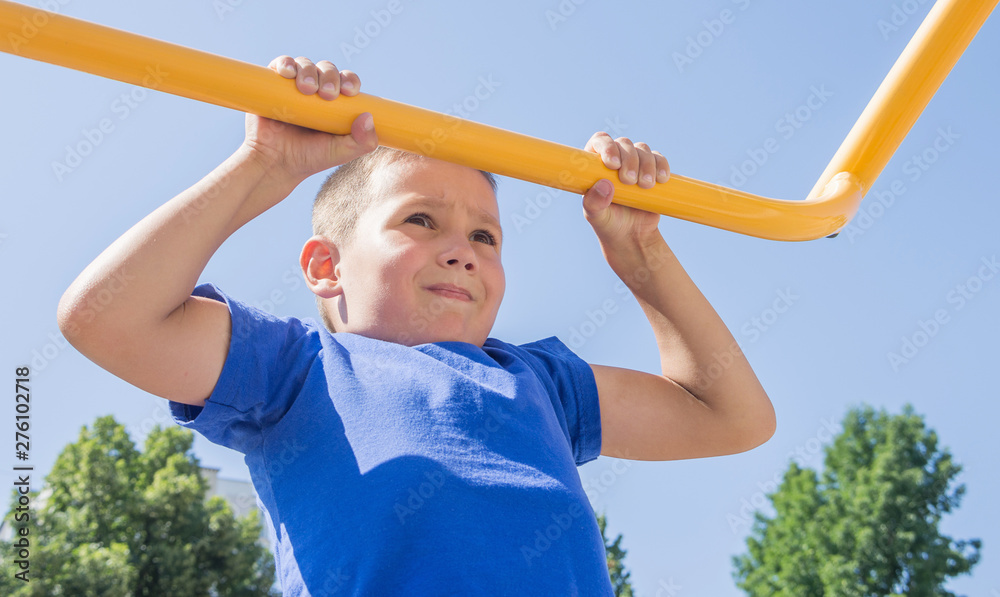 7 year old boy is training to pull up on the horizontal bar. Emotions on the face of a child from difficult sports exercises. Healthy lifestyle from childhood, concept. 7 year old boy is training to pull up on the horizontal bar. Emotions on the face of a child from difficult sports exercises. Healthy lifestyle from childhood, concept.