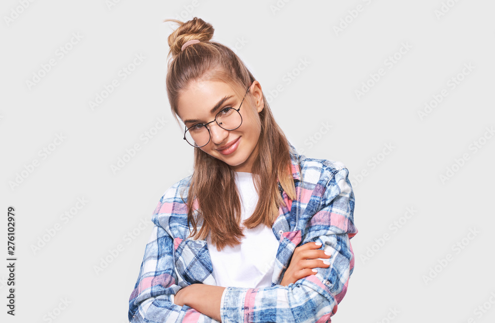 Positive young woman dressed in casual outfit, wearing round transparent eyewear with pleasant smile, keeps both arms crossed, looking to the camera and posing over white studio wall. People emotions Positive young woman dressed in casual outfit, wearing round transparent eyewear with pleasant smile, keeps both arms crossed, looking to the camera and posing over white studio wall. People emotions