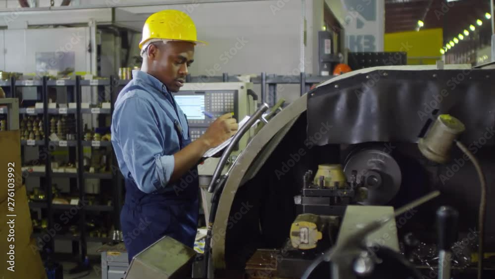 Thigh-up shot of dark-skinned lathe operator standing in workshop and filling out paperwork on clipboard, glancing at control panels, wearing blue overalls and hardhat, with shelves of produce at back Thigh-up shot of dark-skinned lathe operator standing in workshop and filling out paperwork on clipboard, glancing at control panels, wearing blue overalls and hardhat, with shelves of produce at back
