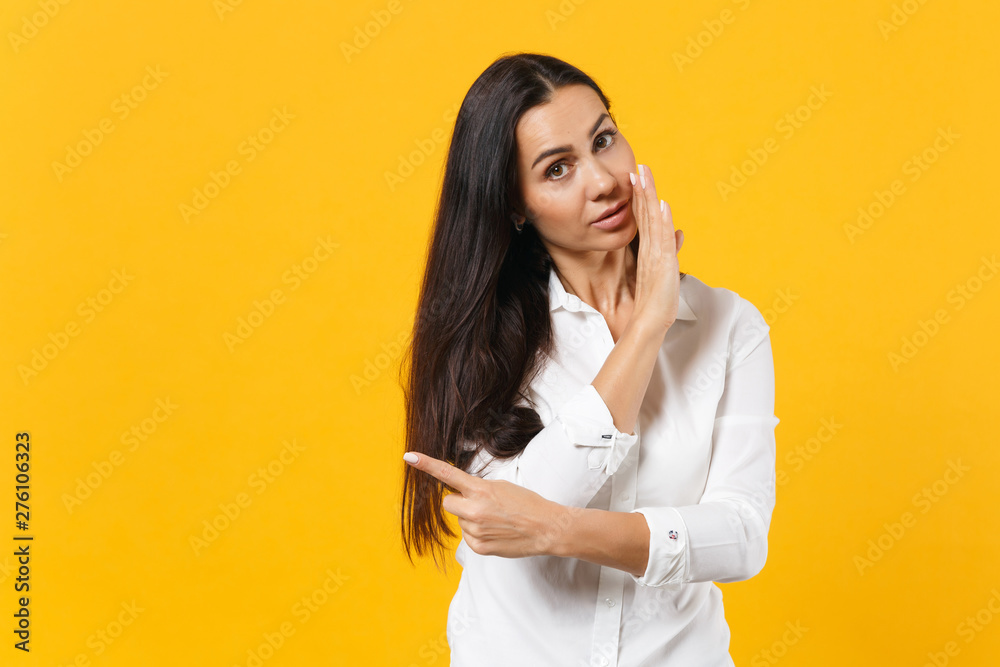 Portrait of young woman in white shirt whispers gossip, tells secret with hand gesture, pointing index finger aside isolated on yellow orange background. People lifestyle concept. Mock up copy space. Portrait of young woman in white shirt whispers gossip, tells secret with hand gesture, pointing index finger aside isolated on yellow orange background. People lifestyle concept. Mock up copy space.