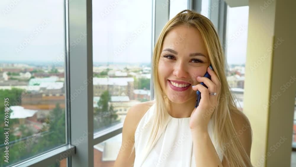 happy woman looks at camera and talks on phone with cityscape behind window happy woman looks at camera and talks on phone with cityscape behind window