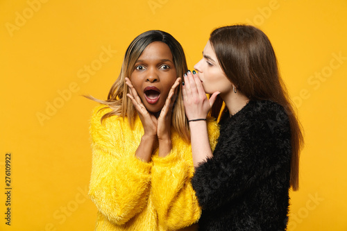 Two young women friends european and african american in black yellow clothes standing posing isolated on bright orange wall background, studio portrait. People lifestyle concept. Mock up copy space.