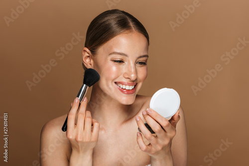 Amazing woman posing isolated over brown chocolate background wall holding mirror apply makeup with brush.