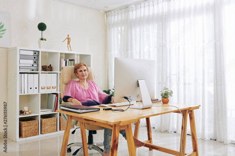 Relaxed smiling aged woman sitting at her table in office of creative studio Relaxed smiling aged woman sitting at her table in office of creative studio
