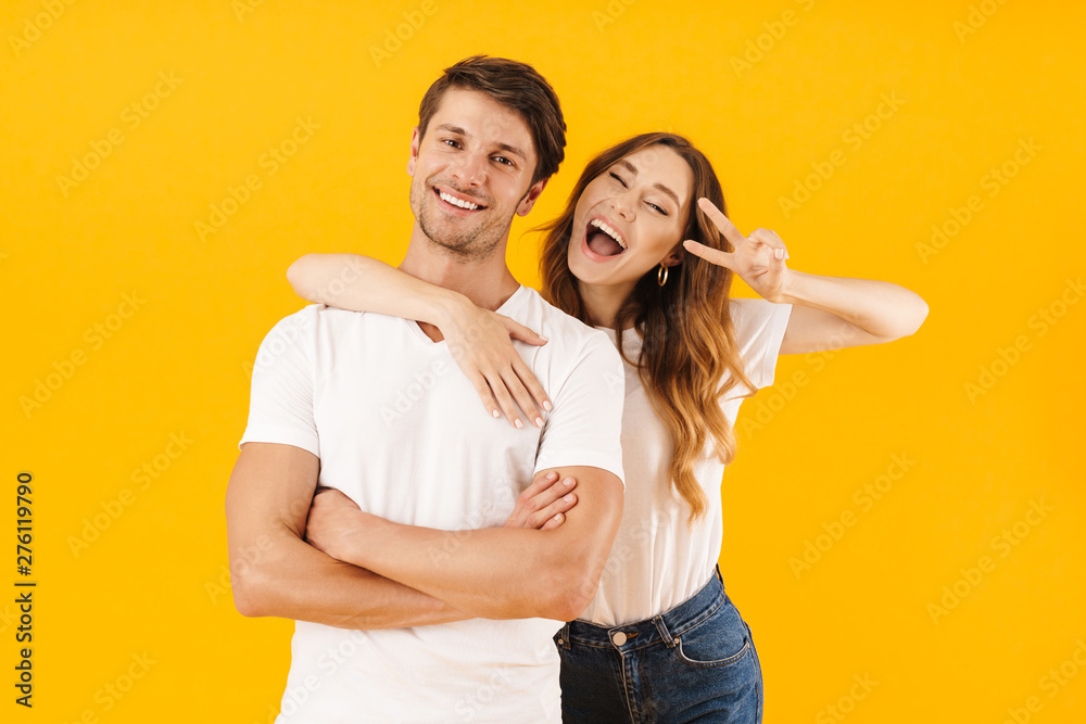 Naklejka premium Portrait of joyous couple in basic t-shirts standing together while woman hugging man and showing peace sign at camera