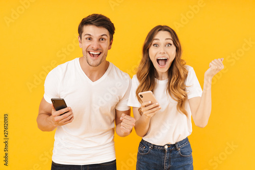 Portrait of happy couple man and woman in basic t-shirts rejoicing while standing together with smartphones