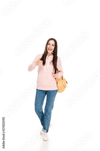 full length view of smiling brunette girl with backpack showing thumb up isolated on white