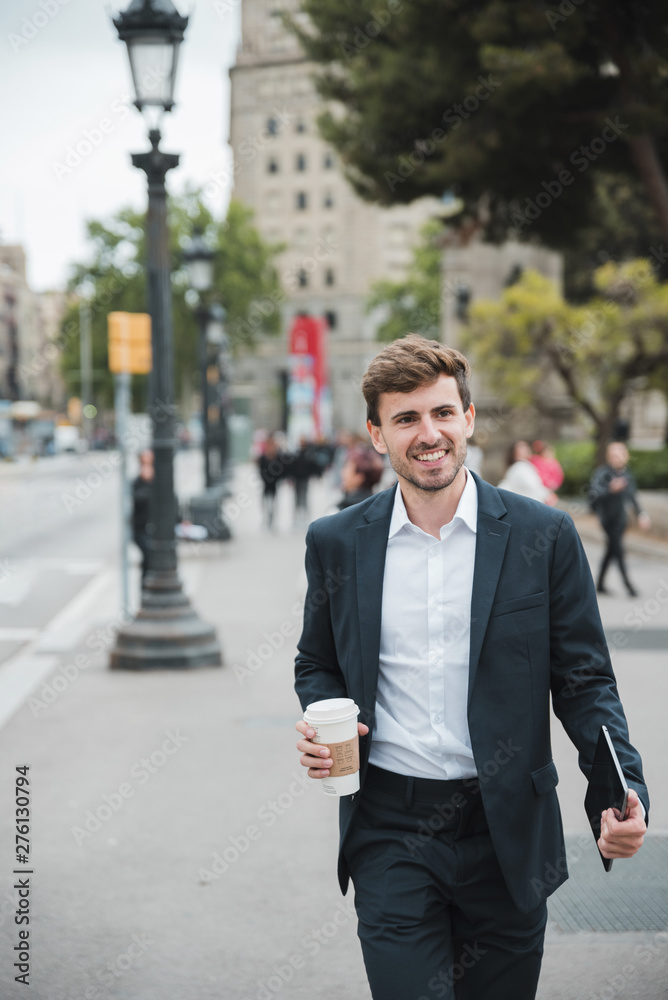 Happy young businessman walking on street with his digital tablet and disposable coffee cup Happy young businessman walking on street with his digital tablet and disposable coffee cup