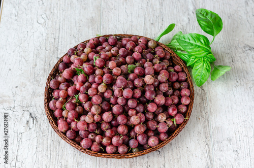 Juicy ripe red berries of a gooseberry in a small wicker round basket and a sprig of fresh basil on a wooden background
