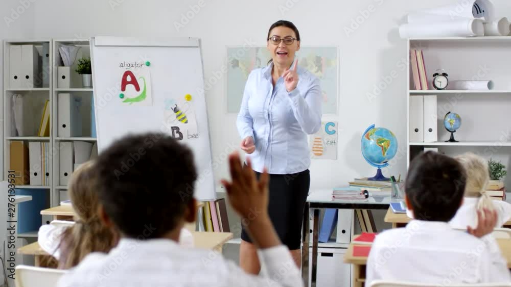 Tracking shot of female primary teacher in glasses standing at whiteboard and teaching alphabet to cute little children sitting at their desks and raising hands to answer questions Tracking shot of female primary teacher in glasses standing at whiteboard and teaching alphabet to cute little children sitting at their desks and raising hands to answer questions
