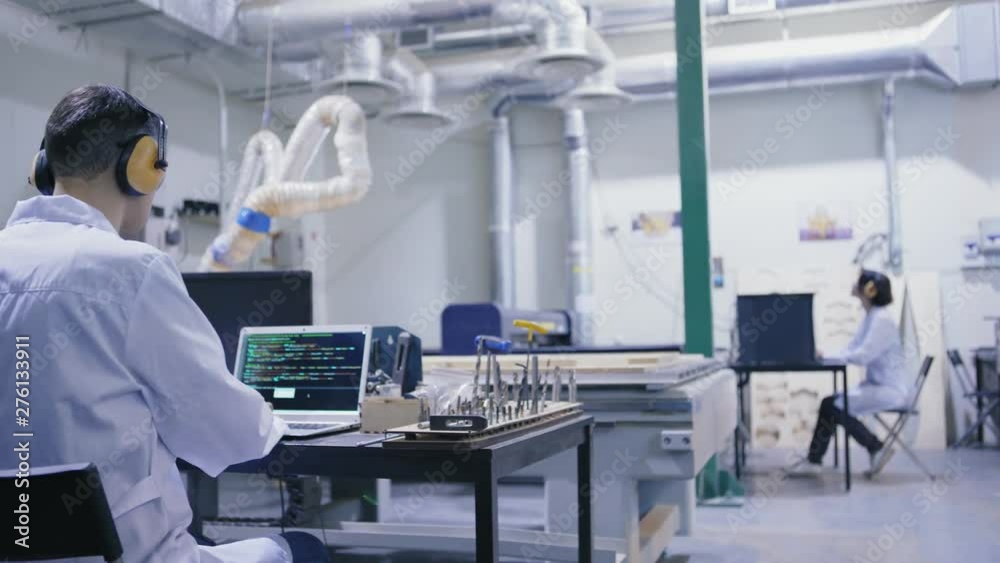 Tracking right back view shot of cnc engineer in white coat and safety ear muffs working on laptop computer in laboratory with laser engraving machines