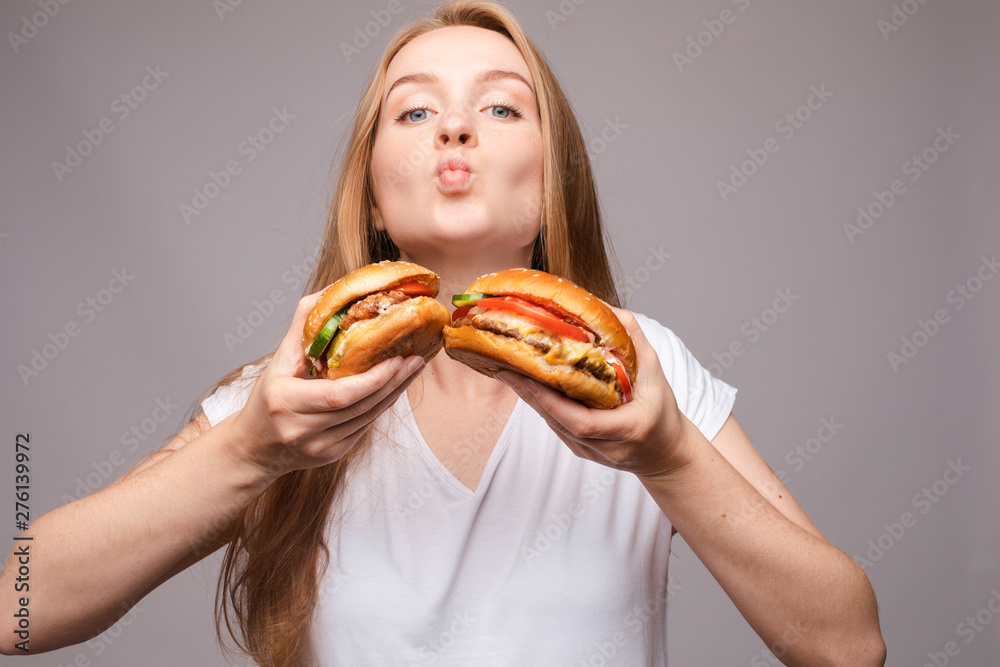 Close-up of pretty long-haired girl biting delicious burger with chicken and salad, looking at camera against pink background. American fast food concept.