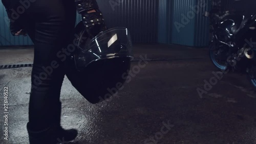 Brutal biker girl carries a glossy black helmet in his hand. The rider approaches his motorbikes in the garage. Cooking in the night races. Moto service.