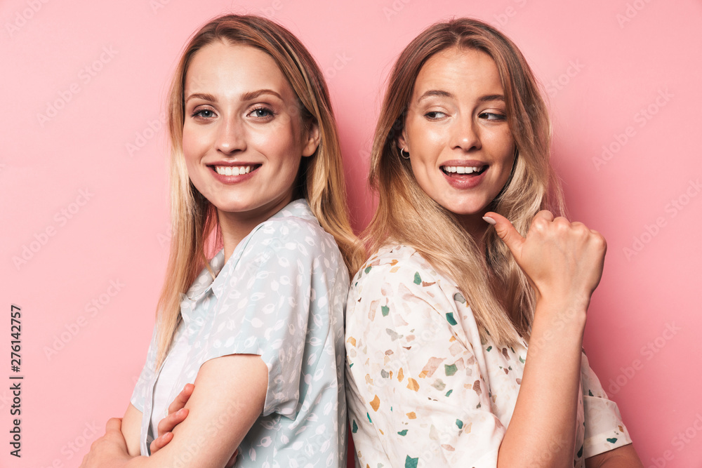 Cheerful woman pointing to her happy pretty blonde women friend sister posing isolated over pink wall background. Cheerful woman pointing to her happy pretty blonde women friend sister posing isolated over pink wall background.